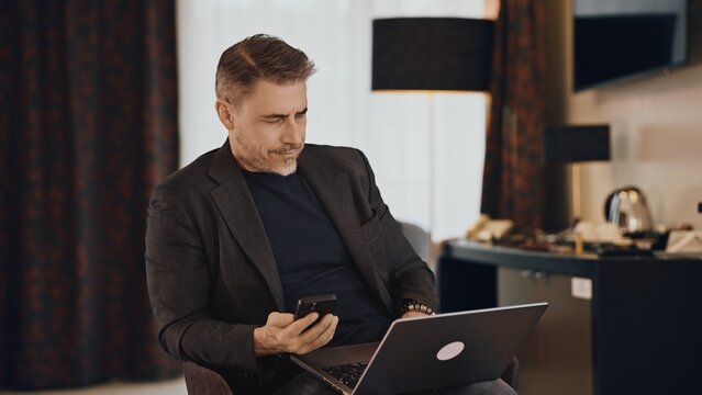 Mature man in a business jacket multitasking with a laptop computer and smartphone in a hotel room. Businessman working on business trip.