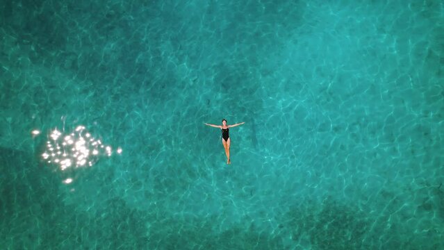 Female gracefully floating on ocean surface. Aerial view of a person spread arms relaxing in crystal clear turquoise sea with sun glimmers. Summer vacations.