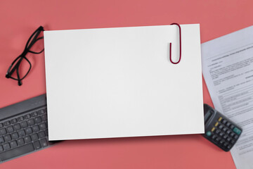 Blank paper sheet with paper clip on blurred background of pink table with a keyboard, glasses,...