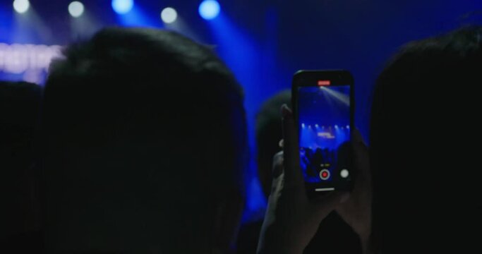 a woman holding a phone during a concert and filming a scene. There is a recording of the concert on stage on the phone display. Prohibited filming of an artist's performance on a smartphone