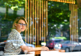 Woman in glasses sitting in cafe or office behind glass with laptop on table. Office work, summer sunny day, freelancing, woman smiling, business project