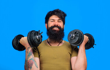 Smiling bearded man with dumbbells up in hands. Strong sportsman doing workout in fitness gym. Athletic muscular man lifting dumbbells in sport club. Weight training and bodybuilding. Sport equipment.