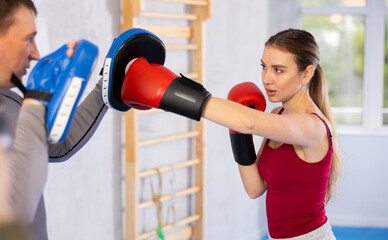 Sportive young girl training boxing kicks on punch mitts held by male instructor in sports hall