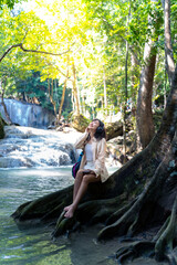 Young Asian woman relax and enjoy outdoor lifestyle travel nature tropical forest on summer holiday vacation. Happy generation z girl listening to the music on headphones and mobile phone by waterfall