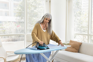 Serious senior Latin maid woman ironing clothing in living room, pressing hot iron to board, using electrical equipment, doing housework, household chores in home apartment