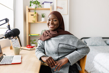 Portrait of young black woman in muslim headscarf smiling at camera sitting at desk in her room. Youth and ethnic people concept. - obrazy, fototapety, plakaty