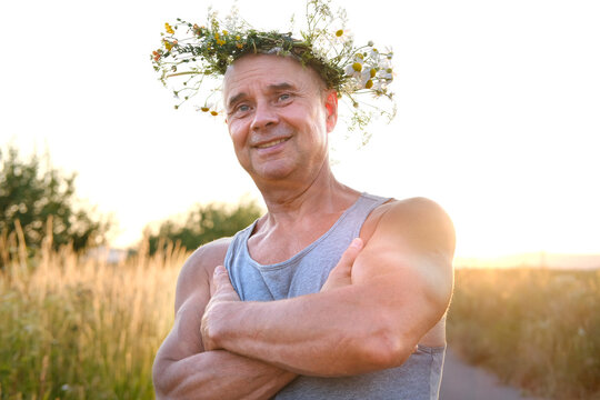 happy cool satisfied muscular funky hipster mature man of 60 years old smiles pretty in T-shirt in sunlight, cute wreath wildflowers, daisies on head, happiness in old age, connection with nature