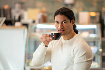 A brunette Hispanic man with long hair drinking coffee from a black mug.