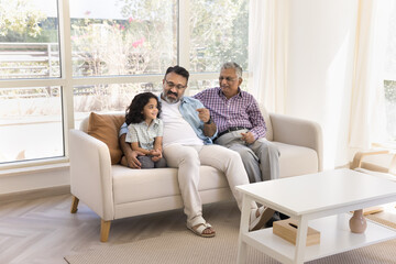 Happy Indian elderly great grandpa and great granddad watching cute little granddaughter kid, enjoying childcare, family leisure, sitting on white sofa together in cozy home living room interior