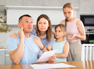 Fototapeta premium Distressed adult man sitting at kitchen table at home and looking worriedly at papers while wife and two teenage daughters trying to calm him down..