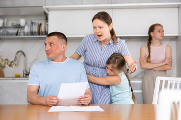 Angry dissatisfied woman standing with tween daughter and chiding upset puzzled man sitting with papers at table in home kitchen