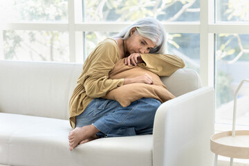 Depressed retired elderly woman sitting on couch at home, keeping closed posture, embracing...