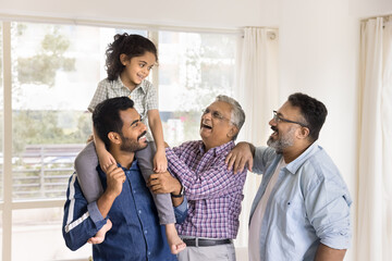 Cheerful Indian daddy, granddad and great grandpa playing with little kid girl sitting on dads shoulders, laughing, standing close together at home, having fun, talking, enjoying family playtime