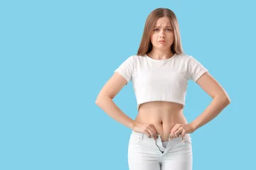 Gardinen Upset young woman trying to button tight pants on blue background. Weight gain concept © Pixel-Shot