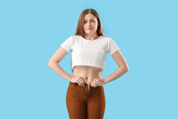 Fototapeta premium Upset young woman trying to button tight pants on blue background. Weight gain concept