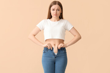 Fototapeta premium Sad young woman in tight jeans on beige background. Weight gain concept