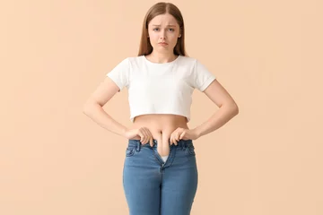 Tapeten Sad young woman in tight jeans on beige background. Weight gain concept © Pixel-Shot