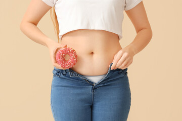 Young woman in tight jeans with doughnut on beige background, closeup. Weight gain concept