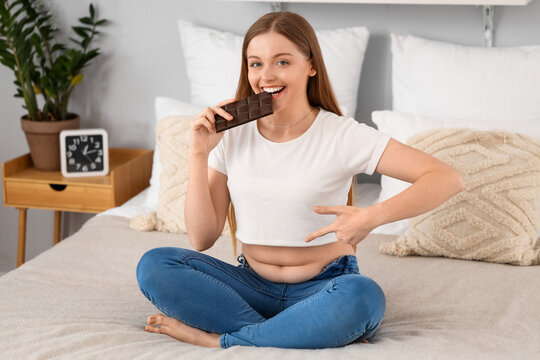 Happy young woman in tight jeans eating chocolate at home. Weight gain concept
