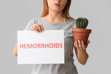 Mature woman holding paper with word HEMORRHOIDS and cactus on light background, closeup