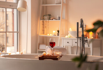 Stand with wine, towels and burning candles on bathtub in room
