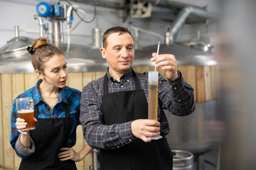 Couple of professional brewmasters standing on background of fermenters at brewery, using test tube...