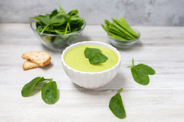 Homemade spinach cream soup in white bowl with bread on white wooden background. 