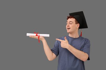 Male student in mortar board pointing at diploma on grey background