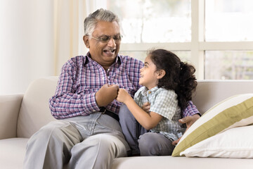 Cheerful old Indian great grandpa and cute little grandkid girl giving fist bumps, talking and laughing on couch, celebrating success, achievement, enjoying friendship, trust, family leisure