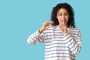 Beautiful young African-American woman with sweet chocolate candy showing silence gesture on blue background