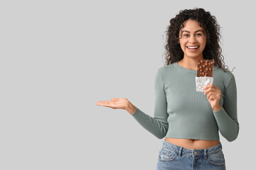Happy young African-American woman with sweet chocolate bar showing something on grey background