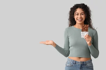 Behang Happy young African-American woman with sweet chocolate bar showing something on grey background © Pixel-Shot