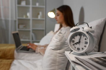 Alarm clock on table of young woman using laptop in bedroom at night, closeup