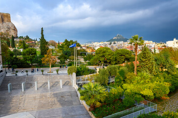 The tree lined Plaka neighborhood under the Acropolis Museum and ancient Acropolis Hill, with Mount Lycabettus covered in rain clouds in the distance, Athens Greece.