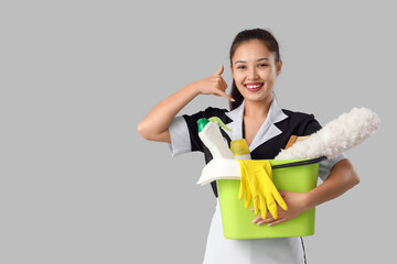 Young Asian chambermaid with cleaning supplies showing "call me" gesture on grey background
