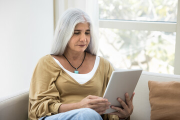 Focused grey haired senior lady using tablet at home, sitting on comfortable couch, reading ebook on screen, watching video content, enjoying retirement, leisure with wireless gadget