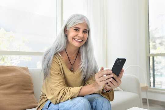 Cheerful positive grey haired senior Latin woman using online application, ecommerce service on mobile phone, posing for home portrait on white couch, looking at camera with toothy smile