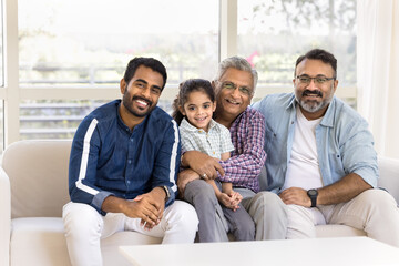 Happy Indian great grandpa holding little kid girl in arms on lap, sitting at mature son and young...