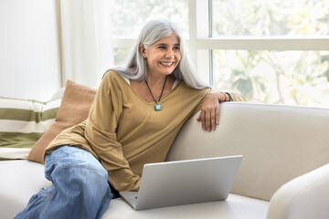 Happy grey haired older Latin woman using laptop at home, resting on couch, typing on computer,...