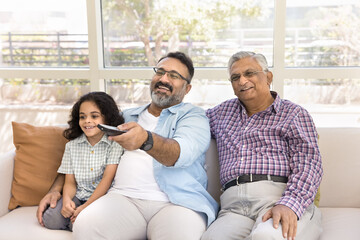 Happy Indian great grandpa, granddad and cute little grandkid girl watching TV at home together,...