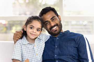 Cheerful handsome Indian dad and beautiful little daughter kid looking at camera, smiling, posing for home headshot portrait. Happy father hugging cute daughter video call screen head shot