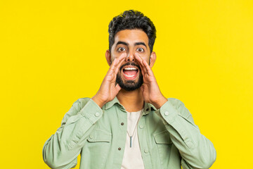 Happy Indian young man yell, shout to camera, promotion advertisement of holidays sale, invitation, greetings, welcome. Excited smiling Arabian guy scream loud isolated on yellow studio background