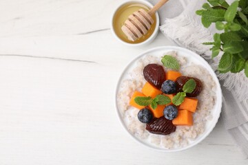 Delicious barley porridge with blueberries, pumpkin, dates and mint served with honey on white table, flat lay. Space for text