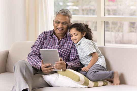 Happy Indian grandfather and little granddaughter girl using tablet for Internet communication, talking on family video call, sitting on home couch together, holding digital device, smiling - Powered by Adobe