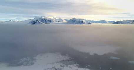 Fototapeta premium Fly over sunset clouds, mountain peak above sunrise fog cloudscape in Antarctica. Ocean aerial view snow covered surface of Antarctic continent. Wild nature background. Drone flight