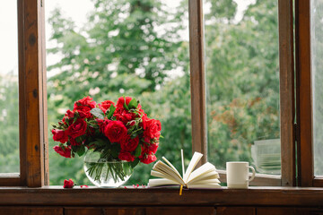Bouquet of red rose in a vase and a book on a wooden window sill. Still life on the window of an old country house, summer cottage. Floral home decoration.