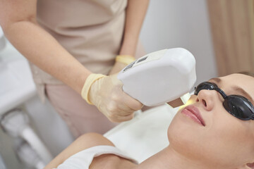 Laser facial treatment for hair removal and rejuvenation at spa. White equipment enhances beauty and skin care. The cosmetologist uses advanced technology for therapy, promoting health, cosmetic care