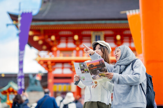 Travel, muslim, Two Asian female tourists of different religions friends visitor learning about history of fushimi inari shrine in travel book while walking through senbon torii path in Kyoto Japan.