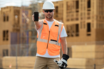 Builder in a hard hat working on a construction project at a site. Worker hold phone, builder show phone screen. Architect with mobile phone. Man worker in builders helmet on the building site. - obrazy, fototapety, plakaty