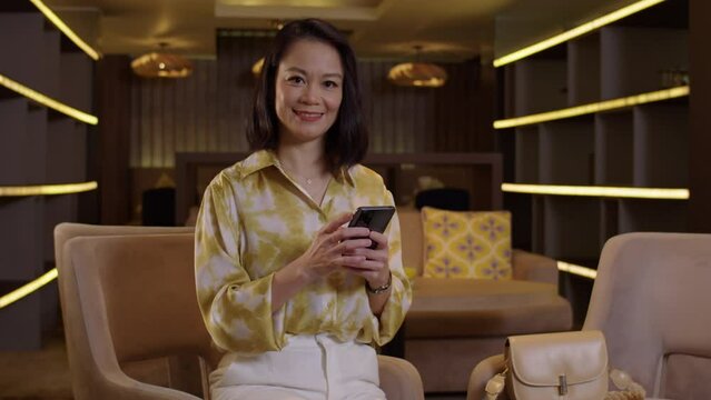 Tilt up portrait of Asian businesswoman checking messages on smartphone sitting at hotel lobby looking at camera